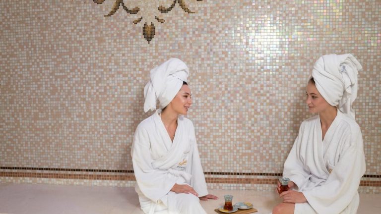 Two women in bathrobes and head towels enjoying a relaxing spa day with Turkish tea indoors.